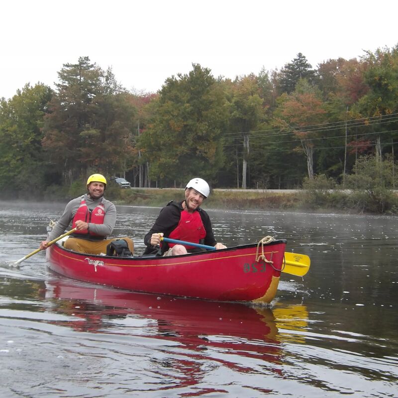 Two men are paddling a red canoe on a river. The man in the front is wearing a white helmet and a red life jacket, while the man in the back is wearing a yellow hat and a red life jacket. A dog is sitting between them. The river is calm, and the trees on the bank are turning color. The sky is overcast.